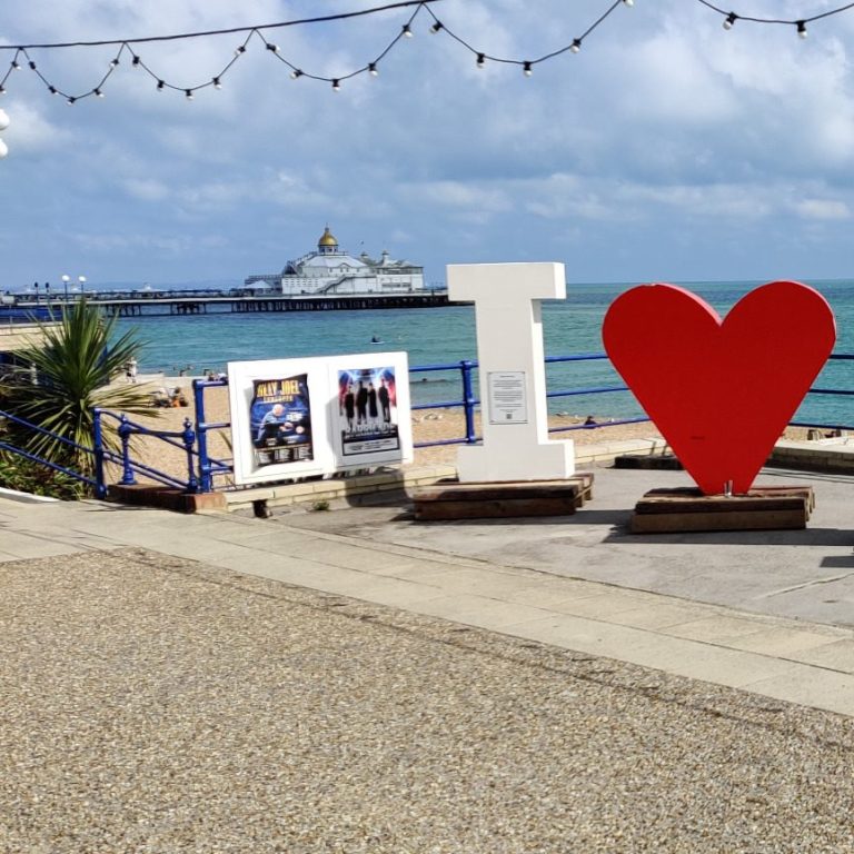 Based in East Sussex Eastbourne Seaside scene featuring large "I ❤️" sign, with pier and sky in the background.