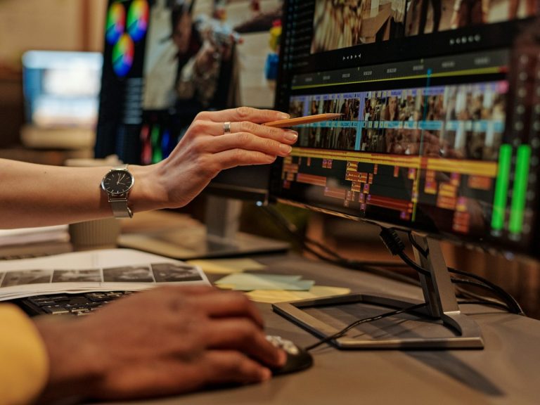 A person indicates a point on the video edit timeline of a computer with multiple screens.