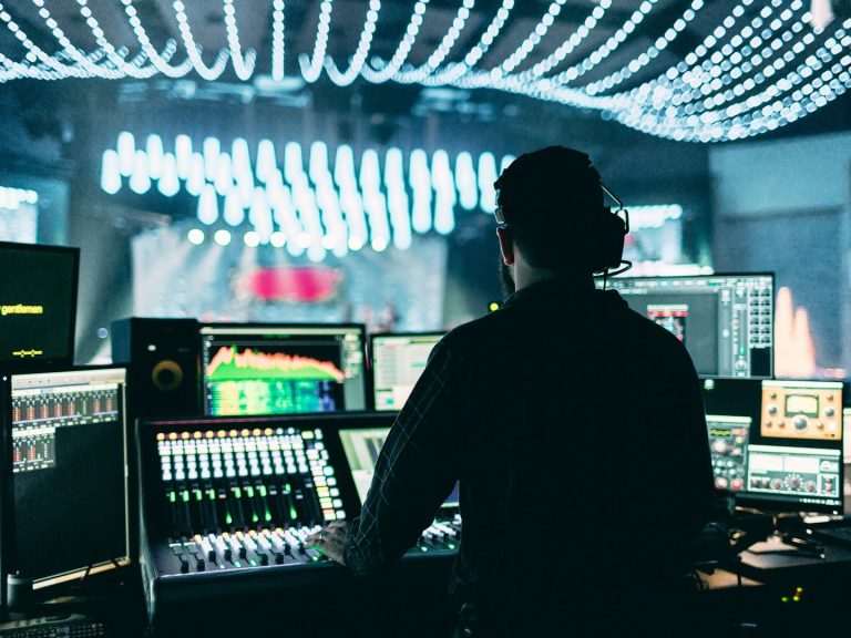 Person operating a sound mixing console in a concert venue with colourful lighting.
