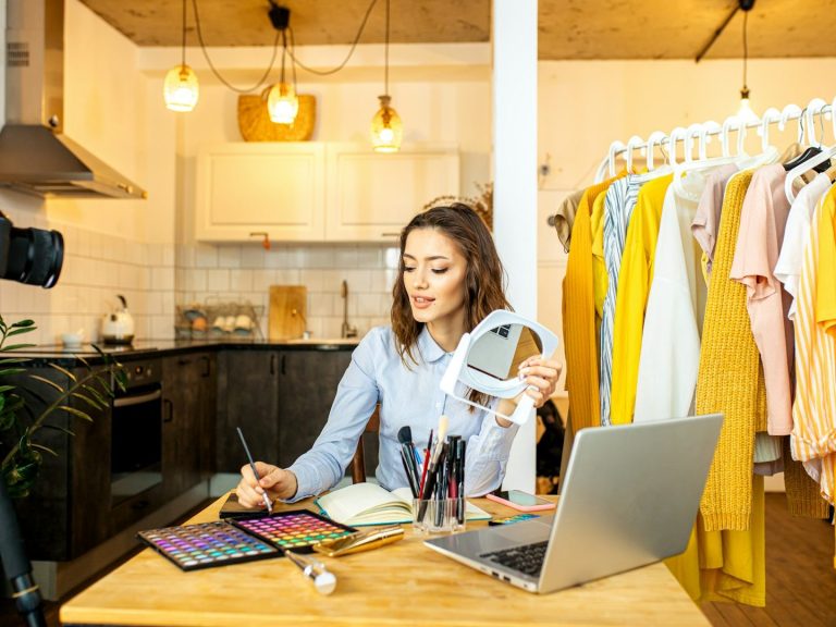 Supporting SME's new to Media Production A woman working at a studio desk with clothes on a rail behind and a laptop, demonstrating make-up application.