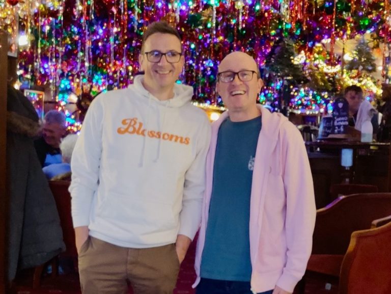 Senior Leadership Gavin and Brian smiling in a pub surrounded by hundreds of Christmas decorations and lights.
