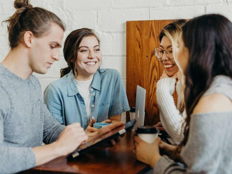 Real World Projects Four people engaged in conversation at an informal workspace, smiling and sharing a tablet.